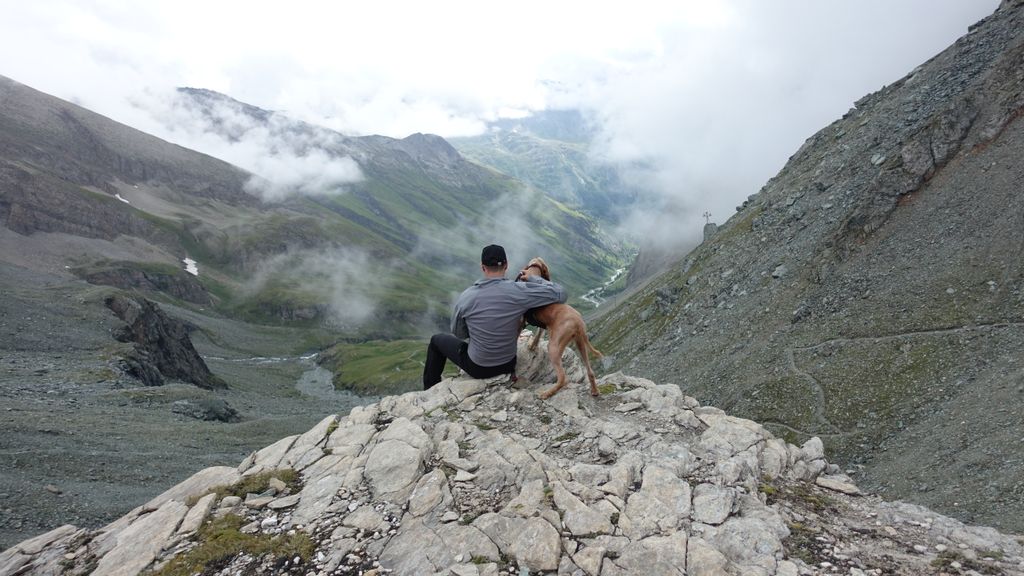 A man and his dog sitting on a rocky peak, overlooking a misty mountain valley.